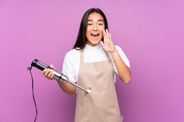 Indische vrouw die handmixer gebruikt die bij purpere muur wordt geïsoleerd die met wijd open mond schreeuwt