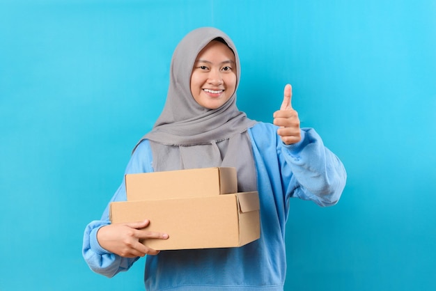 青の背景に分離された親指を示す配達ボックスを保持しているヒジャーブを持つインドネシアのイスラム教徒の女性