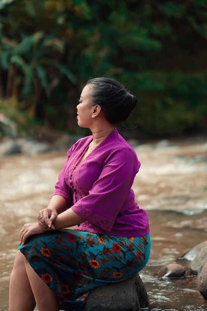 Indonesian woman with purple clothes and makeup on her face sitting near the river with brown color water