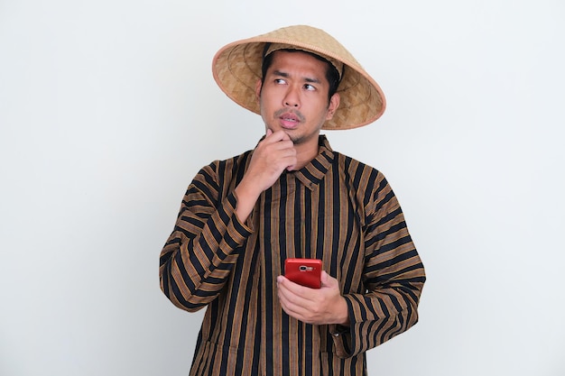 Indonesische traditionele boer die aan iets denkt terwijl hij een mobiele telefoon vasthoudt