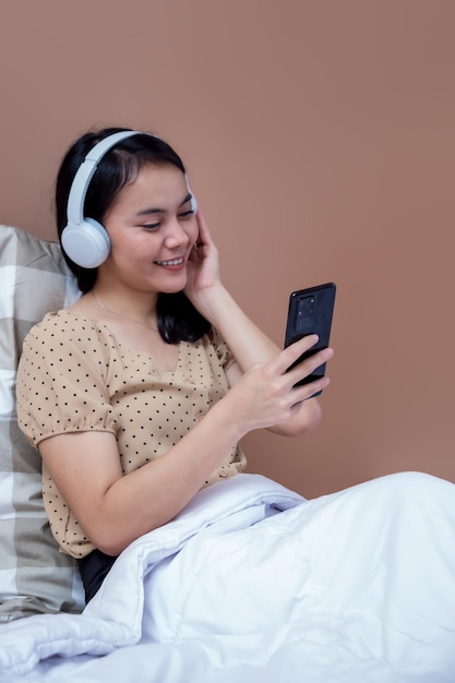 Indonesische vrouw die een koptelefoon draagt en een mobiele telefoon op bed houdt