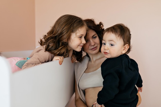 Indoor close up portrait of happy family Woman with her little children is holding them near beds and having dun at home