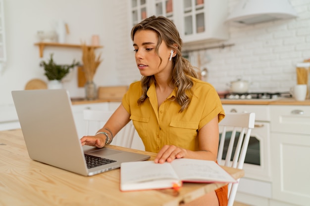 Indoor photo of smiling blond woman using laptop during breackfast on her modern light kitchen.