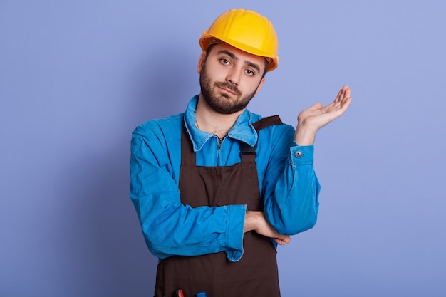 Indoor portrait of concentrated handsome young craftsman looking directly at camera, raising one arm, having hesitation, being disappointed