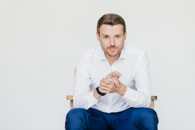 Indoor portrait of handsome self confident businessman in white shirt and black trousers looks with serious expression ready to have business meeting with colleagues isolated over white studio