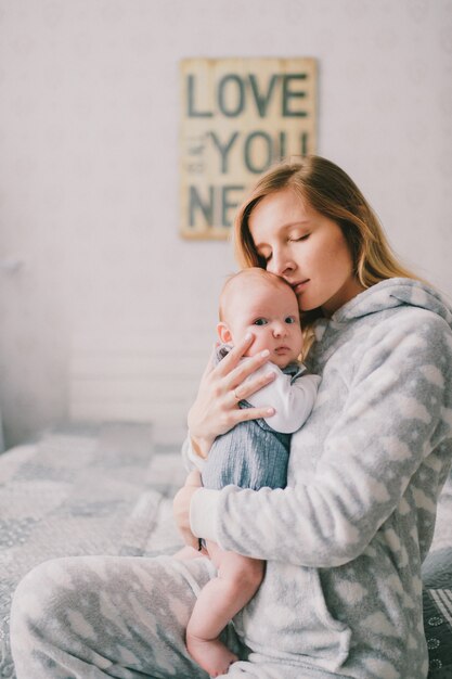 Indoor portrait of  hapy young girl in pijama holding, hugging her newborn baby boy near  wall with decorative plate. Mother take care of her infant child.  Motherhood concept. Love is all you need