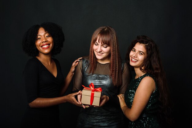 Indoor portrait of three happy bestfriends sisters women, ready for holiday party, holding bright gifts and presents, wearing bright dresses on black background.