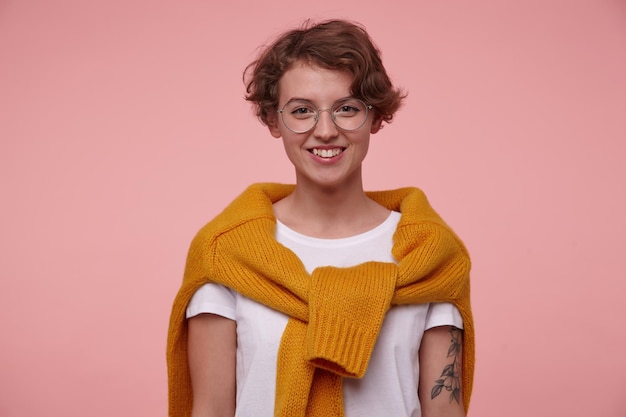 Indoor portrait of young curly female, wears casualty with\
round glasses, smiles broadly, isolated over pink background