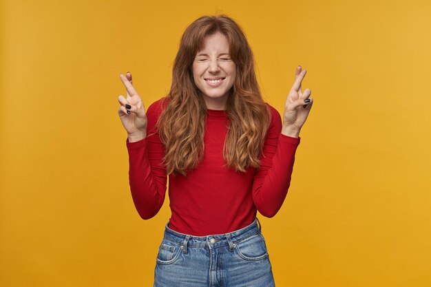 Indoor portrait of young redhead female with wavy long hair keeps her finger crossed, smiling and pray for good exams result, isolated over orange background