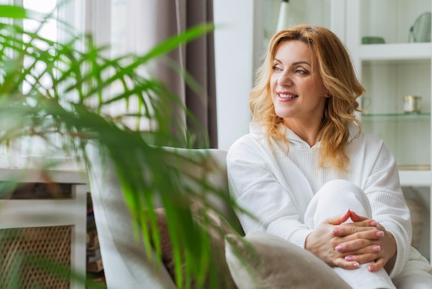 Indoor portret van aantrekkelijke Europese vrouw in haar 40 plus met blond haar en fit lichaam camera kijken met tevreden vriendelijke glimlach, zittend in een witte jurk op comfortabele grijze bank