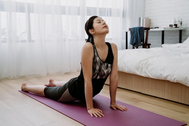 азиатская женщина отдыха в помещении занимается йогой, чтобы снять стресс в спальне. китаянка практикует позу кобры, выпрямляя руки, чтобы поднять грудь с пола, закрывая глаза и делая глубокий вдох