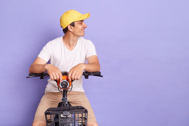 Indoor shoot of young delivery man of takeaway on scooter driving fast express food delivery service caucasian courier on the moto scooter isolated over purple background