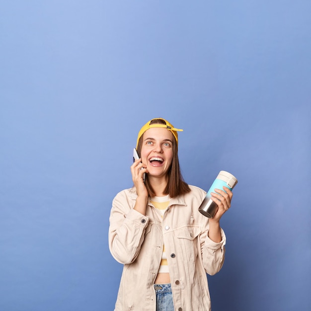 Indoor shot of adorable attractive teenager girl wearing baseball cap and jacket posing isolated over blue background holding thermos and talking phone looking up at copy space for advertisement