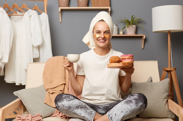 Indoor shot of adorable Caucasian smiling happy woman wearing casual style attire and with towel on head sitting on sofa with clothes hang on shelf on background drinking coffee and eating sweets