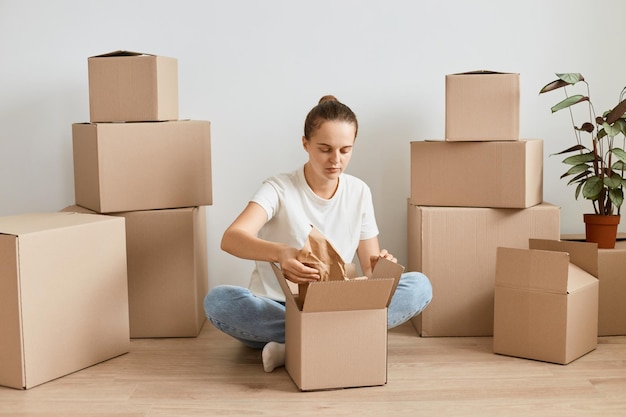 Снимок в помещении очаровательной восхищенной женщины в белой футболке, упаковывающей картонную коробку женщина, упаковывающая вещи, чтобы переехать среди картонных посылок, переезд и переезд в новый дом