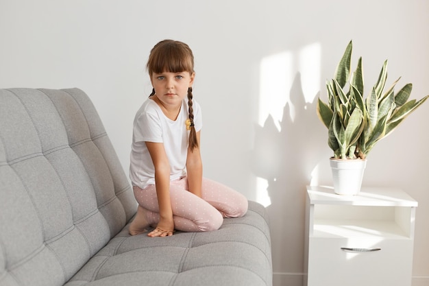 Indoor shot of adorable sad female kid with dark hair and pigtails sitting on couch and looking at camera with upset facial expression, wearing white casual style t shirt.