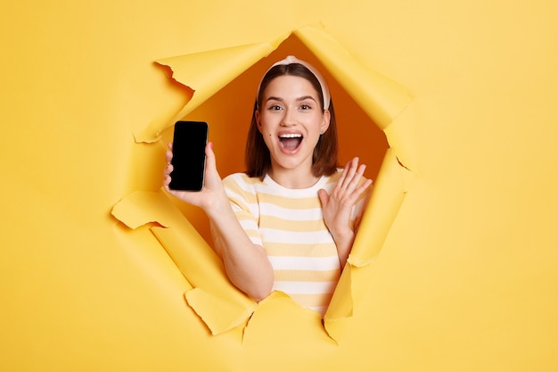 놀란 브루네트 여성의 실내 사진은 찢어진 종이 구멍에 서서 노란색 배경의 돌파구를 통해 광고를 보기 위해 빈 화면이 있는 휴대전화를 보여줍니다.