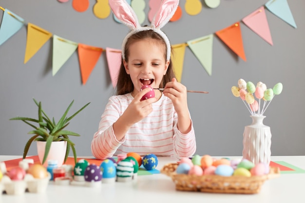 Снимок в помещении изумленной веселой счастливой маленькой девочки, рисующей пасхальные яйца в кроличьих ушах, смотрящей на празднование Пасхи, наслаждающейся процессом подготовки к празднику