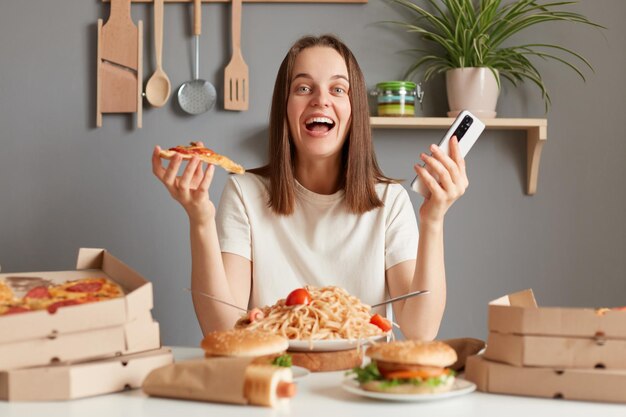 Photo indoor shot of amazed excited young adult woman with brown hair wearing white t shirt sitting in kitchen at table and looking at camera holding in hands smart phone and slice of pizza