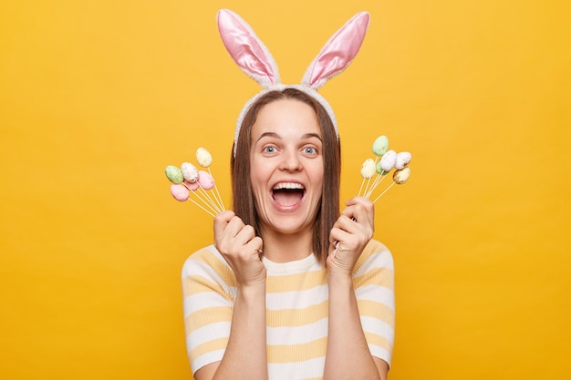 Tiro al coperto di una donna felicissima e stupita vestita di orecchie di coniglio che tiene in mano una torta di pasqua che si apre urlando di felicità festeggiando con uno stato d'animo festoso in posa isolato su sfondo giallo