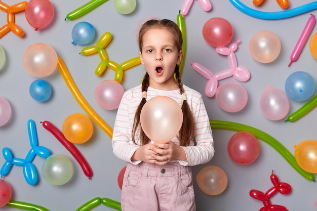 Indoor shot of amazed surprised little girl with braids wearing casual clothing posing isolated over gray background with balloons holding ballon keeps mouth open singing