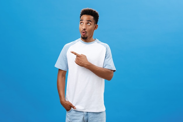 Indoor shot of amazed and thrilled amused handsome dark-skinned guy with beard in casual t-shirt pointing and gazing left with folded lips from excitement and joy saying wow from curious emotions