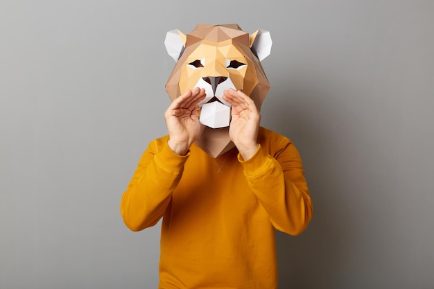 Indoor shot of anonymous man wearing lion mask and orange\
sweatshirt standing with hands near mouth showing his paws\
screaming announcing posing isolated over gray background