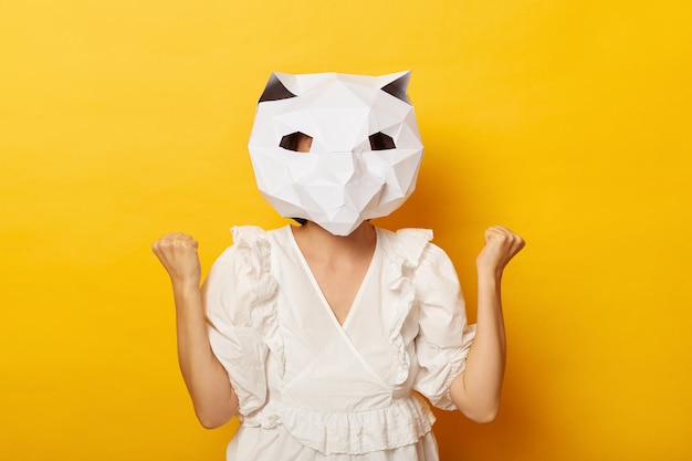 Indoor shot of anonymous woman wearing dress and paper cat mask\
showing her clenched fist celebrating victory posing isolated over\
yellow background