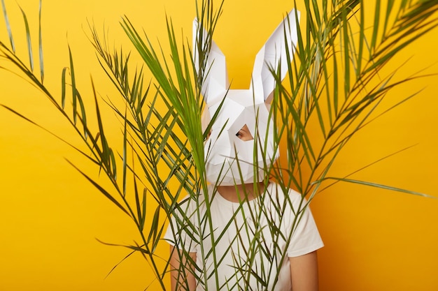 Indoor shot of anonymous woman wearing white Tshirt and paper rabbit mask standing isolated over yellow background hiding behind green palm leaves feels scared