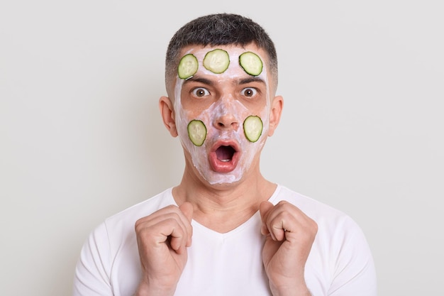 Indoor shot of astonished shocked man wearing white t shirt standing looking at camera with mask and slices of cucumbers being surprised of result posing isolated over gray background