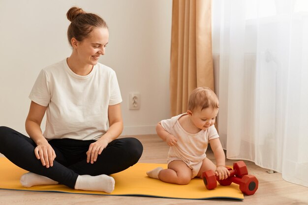 Indoor shot of athletic woman wearing white t shirt and black leggins sitting on floor on yoga mat with her toddler daughter looking at baby with happy smile fitness at home