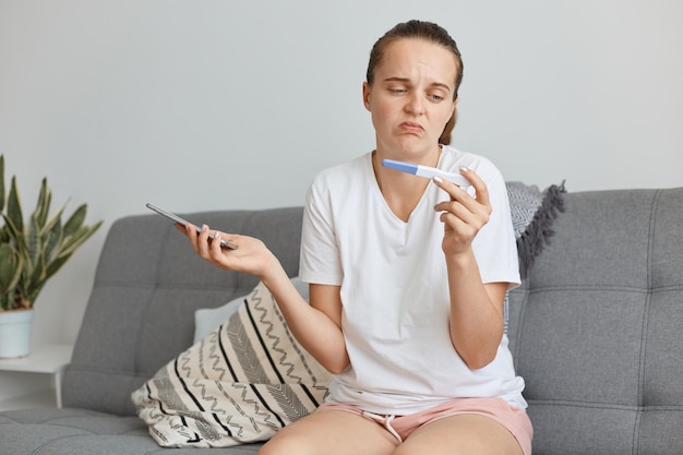 Indoor shot of attractive dark haired woman wearing white t shirt and short, sitting on sofa in living room with pregnancy test with positive result, does not know what to do.