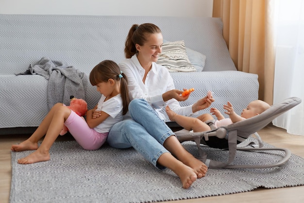 Indoor shot of attractive young woman wearing white shirt and jeans sitting on floor with her daughters elder child posing backwards to mother being offended that all mom's attention to tiny baby