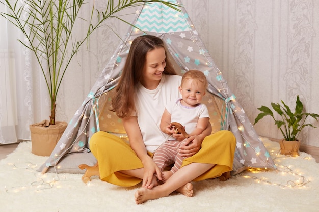 Indoor shot of baby girl and her mother playing at home in wigwam while sitting on the floor child sitting on mommy's legs and holding wooden toy mom looking at daughter with smile