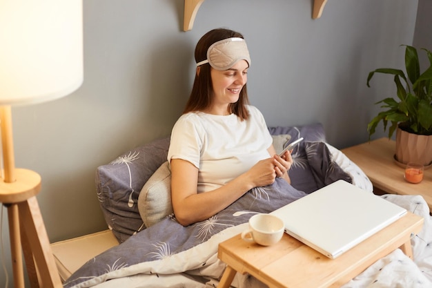 Indoor shot of beautiful brown haired young adult woman wearing\
sleep mask sitting with closed laptop in bed at home finishing her\
online work using smart phone browsing internet