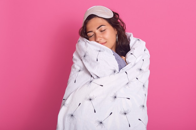 Indoor shot of beautiful brunette caucasian girl wearing white blanket and blindfold on head, woman keeps eyes closed, posing early in morning, isolatedon pink studio. People concept.