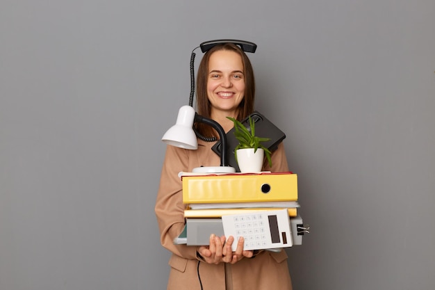 Indoor shot of beautiful smiling attractive funny woman wearing beige jacket with phone on her head holding paper folders posing isolated over gray background having fun at work