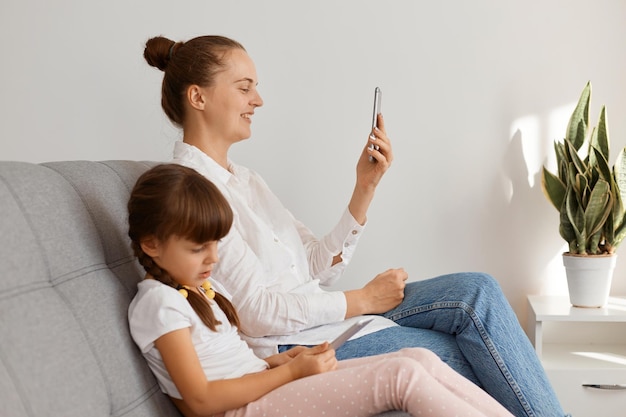 Premium Photo | Indoor shot of beautiful smiling woman with bun hairstyle  wearing white shirt and jeans sitting on sofa with her daughter, female  having video call while kid choosing game to