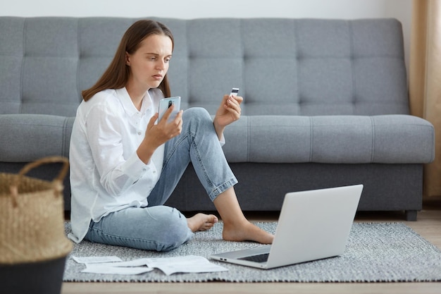 Indoor shot of beautiful woman wearing white shirt and jeans sitting on floor near sofa using laptop holding credit card and smart phone looking with serious and concentrated look at device