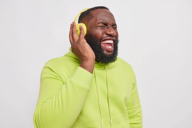 Indoor shot of black bearded man shouts as listens music with very loud sound dressed in hoodie poses against grey studio wall