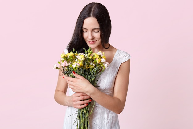 Indoor shot of brunette woman looks gently at flowers in her hands, dressed in white dress, has present from her boyfriend, posing isolated on rosy. Copy space for promotion content.