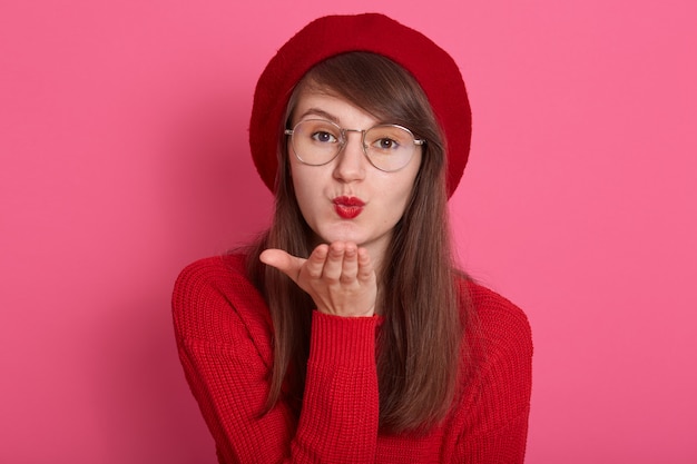 Indoor shot of brunette young Caucasian female blows air kiss, looking directly at camera, expresses her affection, dressed red sweater and beret, has red lips, posing isolated over pink