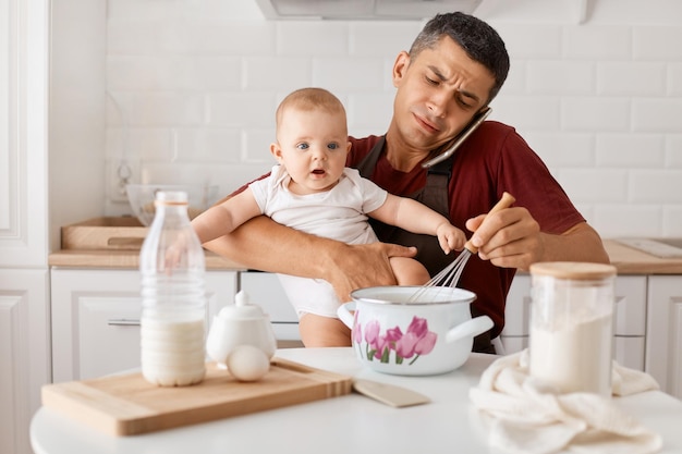 Indoor shot of busy father sitting at table with baby daughter in hands and cooking in kitchen talking phone with sad expression asking how to cook