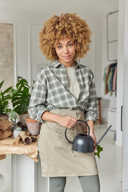 Indoor shot of busy young woman with curly hair holds watering\
can poses in cozy room near table with pots and soil takes care of\
houseplants going to water home flowers home gardening concept