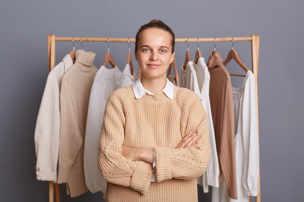 Indoor shot of calm confident woman with bun hairstyle wearing beige jumper standing against gray wall with clothes on hangers on shelf keeps hand folded looking at camera