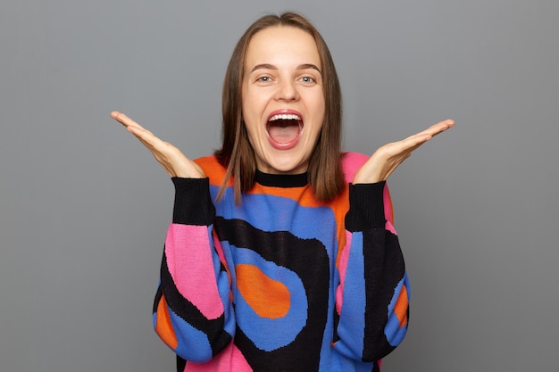 Indoor shot of carefree optimistic woman with brown hair wearing sweater standing isolated over gray background raised her arms yelling with overjoyed expression hearing very good news rejoice