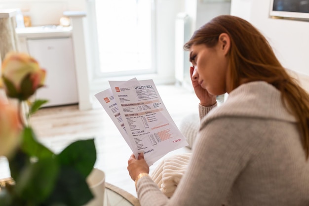 Indoor shot of casually dressed young woman holding papers in her hands, calculating family budget, trying to save some money , having stressed and concentrated look