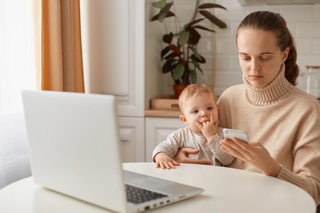 베이지색 스웨터를 입은 백인 검은 머리 여성의 실내 사진은 아기 소녀와 함께 테이블에 앉아 휴대전화를 손에 들고 있다