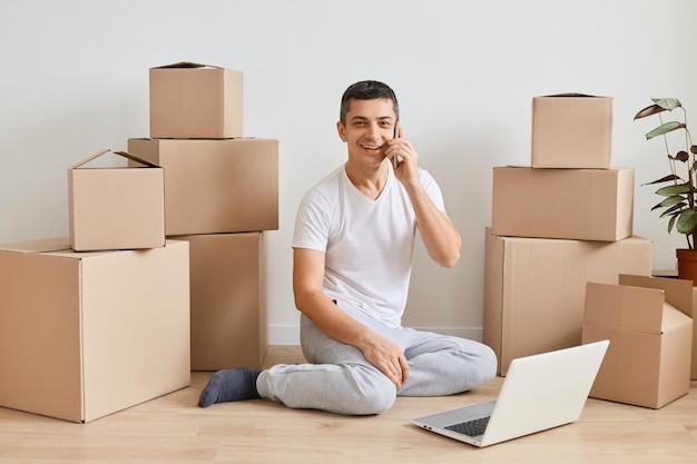 Indoor shot of Caucasian man sitting on floor with laptop before to leave apartment guy talking on mobile phone among cardboard boxes ordering delivery looking at camera