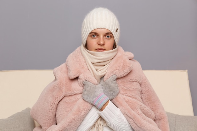 Indoor shot of Caucasian woman wearing cap gloves scarf and coat sitting in living room fells cold trying to warm up waiting for start of the heating season